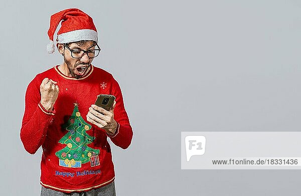 Aufgeregter Mann in Weihnachtsmütze schaut auf Handy und feiert. Menschen in santa Hut Blick auf Handy aufgeregt  aufgeregt junger Mann in Weihnachten Hut mit Handy feiern