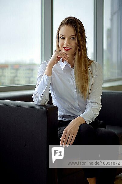 Beautiful and positive businesswoman on the couch looking straight and smiling  window with cityscape on background