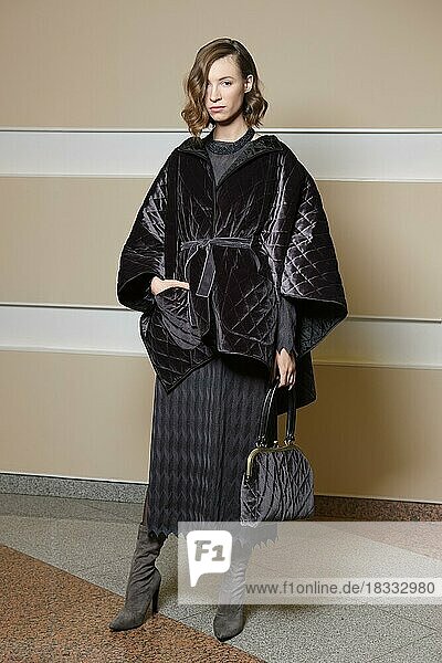 Full Length Porträt des schönen Mädchens in Strickkleid und Poncho Mantel. Wunderschöne Dame in warmen Outfit mit Handtasche posiert in der Nähe der Wand
