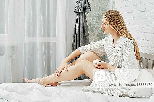 Hübsche Frau trägt Aftershave-Creme auf ihr Bein auf dem Bett sitzend im Profil auf