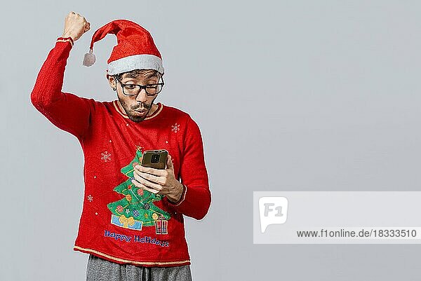 Menschen mit Weihnachtsmannmütze schauen aufgeregt auf ihr Handy  Aufgeregter junger Mann mit Weihnachtsmütze benutzt sein Handy beim Feiern  Aufgeregter Mann mit Weihnachtsmütze schaut auf sein Handy und feiert