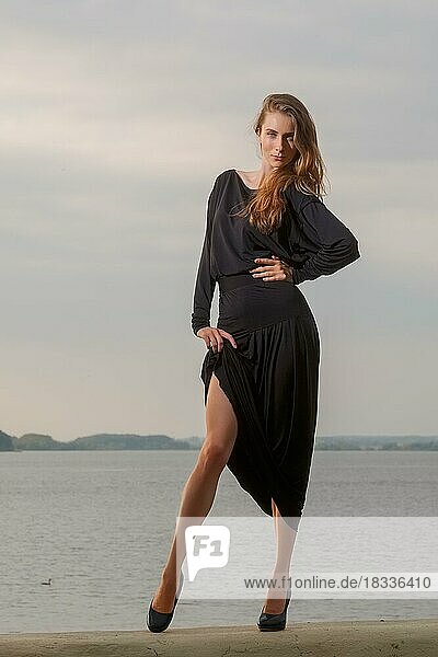 Attraktives Mädchen hebt langes schwarzes Kleid und zeigt ihre schlanken Beine auf dem Pier