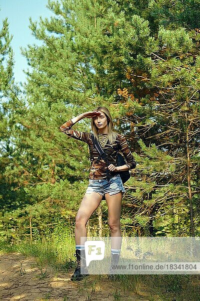 Ganzkörperporträt eines attraktiven Mädchens in Tarnhemd und Jeans-Shorts mit Rucksack auf einem Wanderweg im Wald