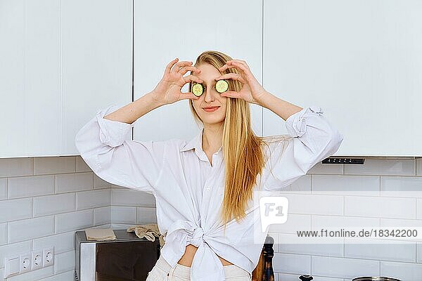 Fröhliche junge Frau hält sich beim Kochen Gurkenstücke an die Augen