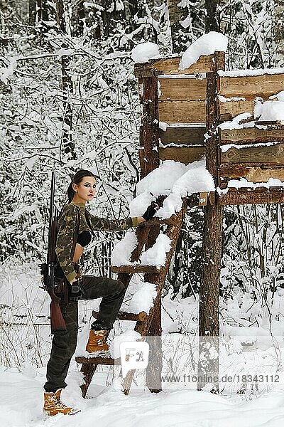 Attraktives Jägermädchen mit Gewehr auf Jagdturm im Winterwald