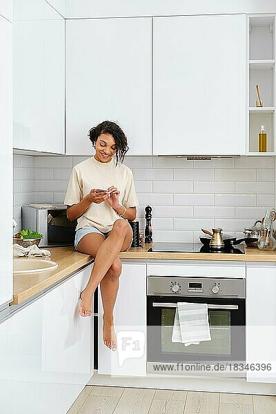 Young woman sitting on kitchen table  reading mobile phone and waiting for the coffee to be brewed on the stove