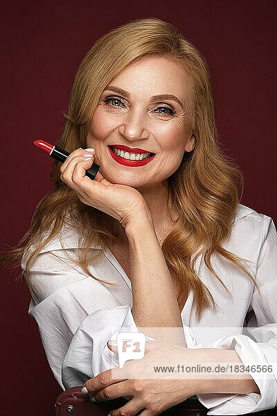 Porträt einer schönen älteren Frau in einem weißen Hemd mit klassischem Make-up und blondem Haar mit rotem Lippenstift in der Hand