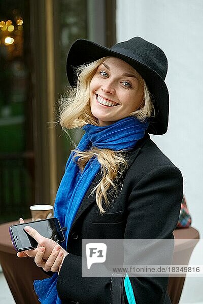 Porträt einer hübschen  fröhlichen Dame in schwarzem Mantel  Hut mit Pappbecher und Telefon in der Hand