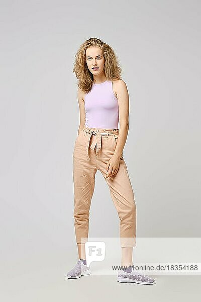 Pretty girl with blonde messy hair in tank top and trousers posing over gray background