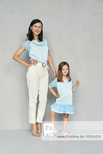 Stylish and trendy mother and her young daughter posing together near gray wall