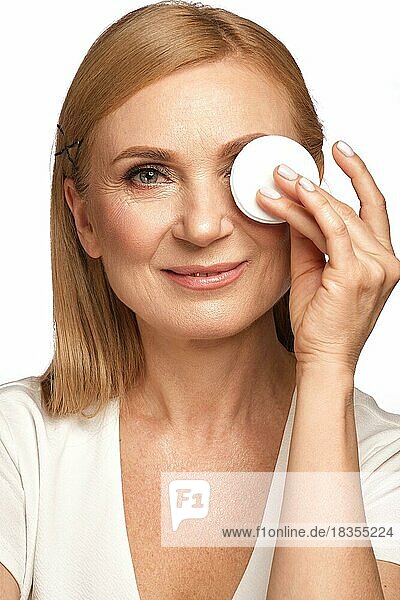 Porträt einer schönen älteren Frau in einem weißen Hemd  die sich mit einem Reinigungsmittel und einem Wattepad abschminken will