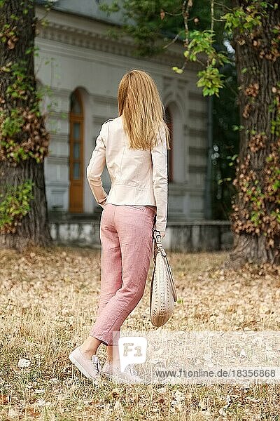 Unrecognizable girl in linen trousers and beige leather jacket walking on ground covered with fallen leaves