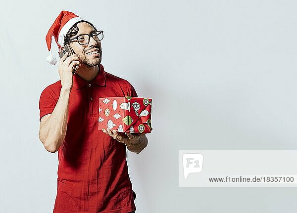 Lächelnder Mann mit Weihnachtsmütze hält Weihnachtsgeschenkbox und spricht am Telefon. Weihnachten Mann hält Geschenk und ruft Familie am Telefon. Glücklicher Mann hält Geschenk und ruft Familie an Weihnachten