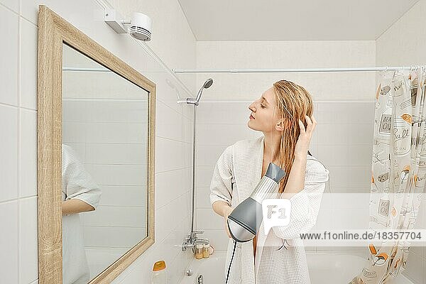 Frau im Bademantel  die ihr Haar mit einem Fön im Profil vor dem Spiegel im Badezimmer trocknet