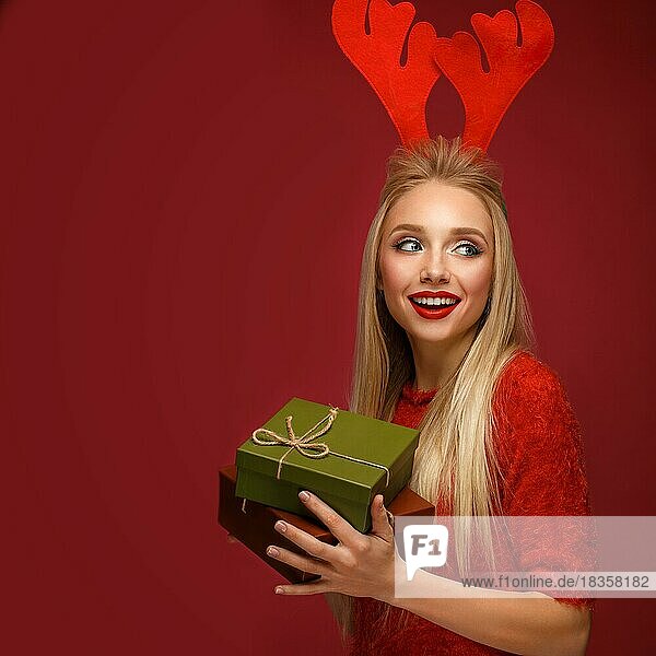 Schönes blondes Mädchen in einem Neujahrsbild mit Kisten mit Geschenken in den Händen und Hirschhörnern auf dem Kopf. Schönes Gesicht mit festlichem Make-up. Foto im Studio aufgenommen