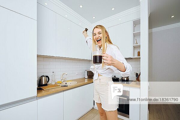 Fröhliche Frau tanzt und singt mit einer Tasse Kaffee in der Hand in der Küche