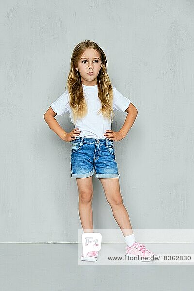 Kleines Mädchen in weißem T-Shirt  Jeans-Shorts und rosa Turnschuhen steht mit den Händen auf der Taille in der Nähe der Wand
