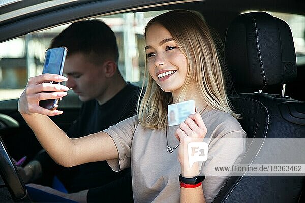 Glücklich lächelndes Mädchen macht ein Selfie mit einem neuen Führerschein  neben einem Fahrlehrer sitzend