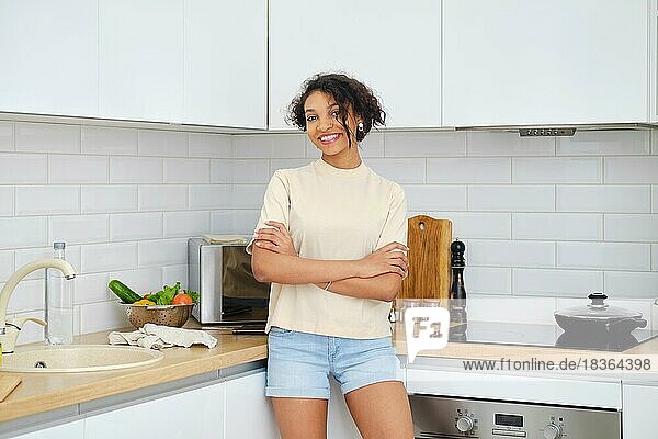 Junge lächelnde afroamerikanische Frau steht in der Küche mit gekreuzten Händen