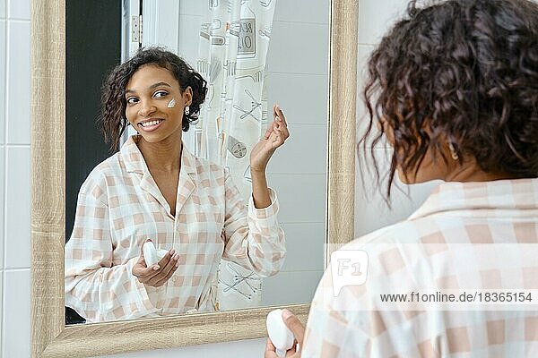 Lächelnde afroamerikanische Frau schaut in den Spiegel und trägt Gesichtscreme im Badezimmer auf