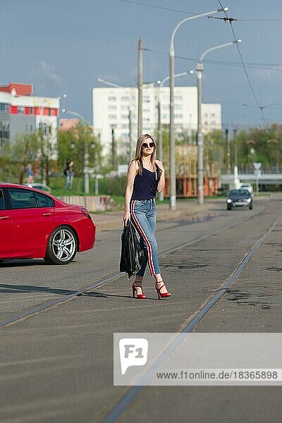 Nettes blondes Mädchen mit Sonnenbrille überquert die Straße mit Lederjacke in der Hand