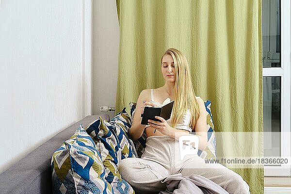 Junge Frau macht sich Notizen in einem Notizbuch und sitzt zu Hause auf der Couch