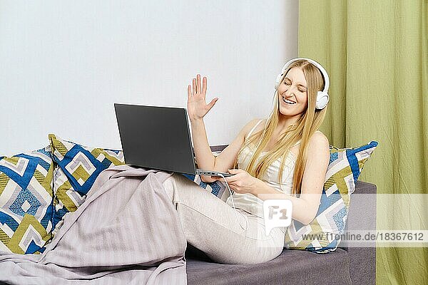 Positive woman on couch waving hand while on video call using laptop and earphones at home during a pandemic