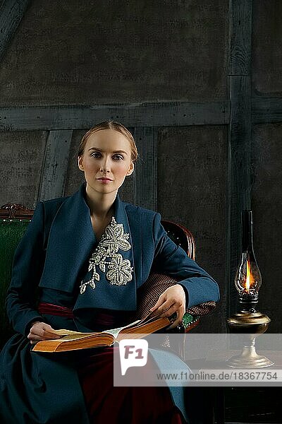 Attractive lady in oldfashioned clothes reading ancient book near vintage kerosene lamp
