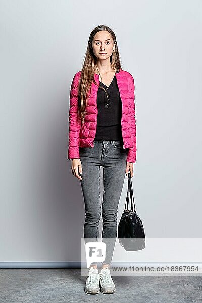 Ganzkörperporträt eines hübschen Mädchens in Daunenjacke und Jeans  das mit einer Handtasche an der Wand steht
