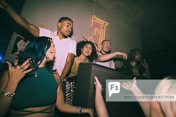 Cheerful young multiracial men and women dancing around DJ at illuminated nightclub