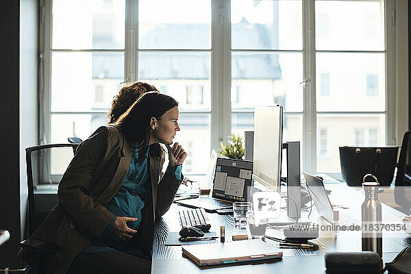 Schwangere Geschäftsfrau mit Hand am Kinn  die auf einen Desktop-PC am Schreibtisch im Büro schaut
