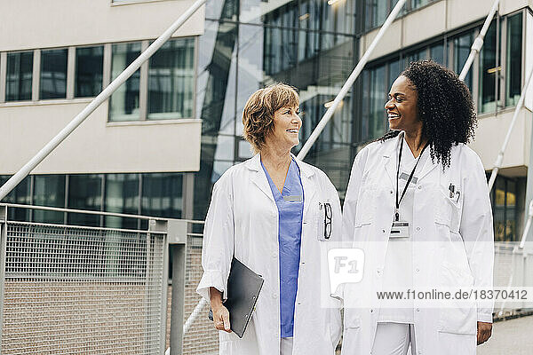 Happy female doctors looking at each other while standing in front of hospital