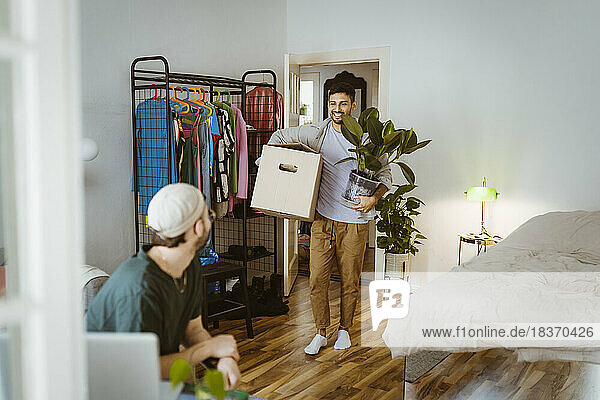Lächelnder Mann  der einen Karton und eine Pflanze hält  während er mit seinem Freund im Schlafzimmer zu Hause spricht