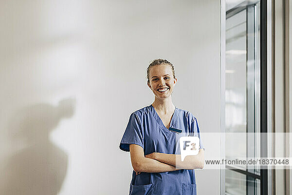 Portrait of happy young female nurse standing with arms crossed against wall at hospital