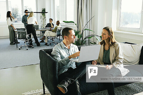 Geschäftsmann erklärt einer Kollegin den Laptop  während er auf dem Sofa im Büro sitzt