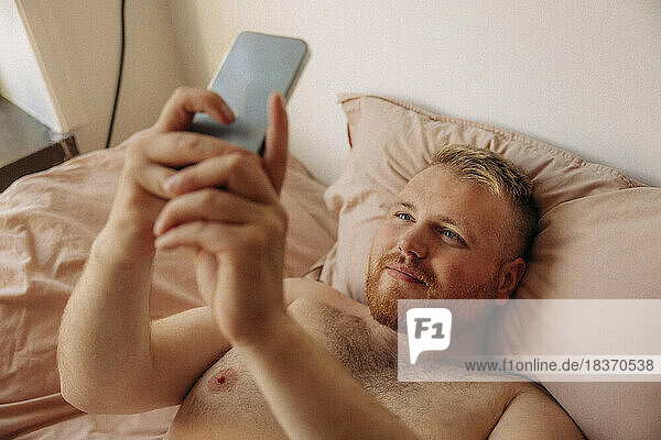 Shirtless obese man taking selfie through smart phone while lying on bed at home
