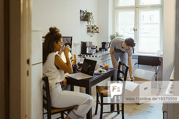 Mann beim Auspacken eines Kartons in der Küche mit seiner Freundin  die zu Hause auf dem Esstisch sitzt