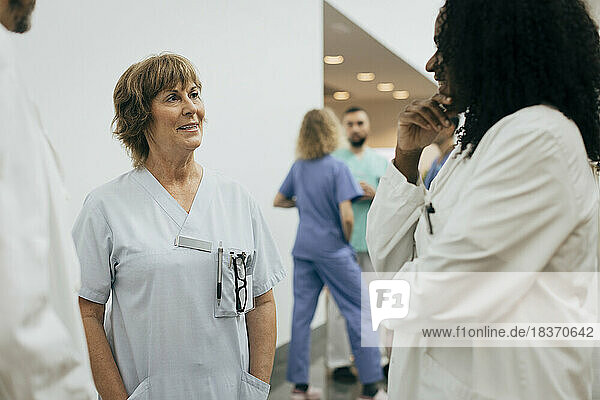 Lächelnde Ärztin mit Händen in den Taschen im Gespräch mit einem Kollegen im Krankenhaus