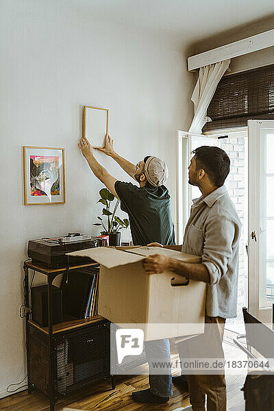 Seitenansicht eines Mannes mit Karton  der bei seinem Freund steht  der zu Hause einen Rahmen an die Wand montiert