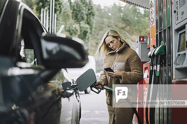 Frau benutzt Mobiltelefon beim Tanken an einer Tankstelle