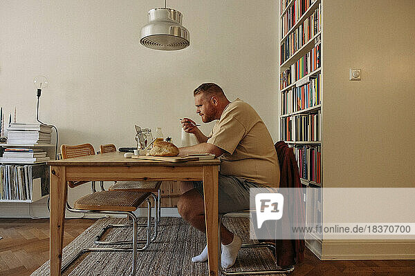 Seitenansicht eines übergewichtigen Mannes  der frühstückt  während er am heimischen Tisch den Laptop betrachtet