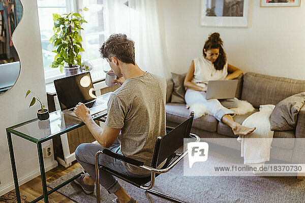 Man having coffee while using laptop sitting on chair near girlfriend in background at home