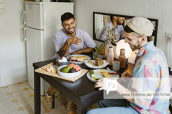 Happy gay couple having fruits while sitting at dining table in kitchen