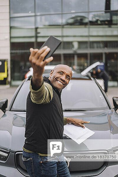 Seitenansicht eines reifen Mannes  der ein Selfie mit einem neuen Auto über sein Mobiltelefon macht