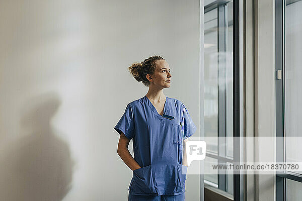 Nachdenkliche junge Krankenschwester steht mit den Händen in den Taschen an der Wand eines Krankenhauses