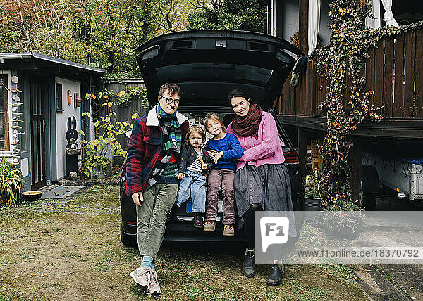 Lächelnde Familie sitzt zusammen im Kofferraum eines Autos vor dem Haus