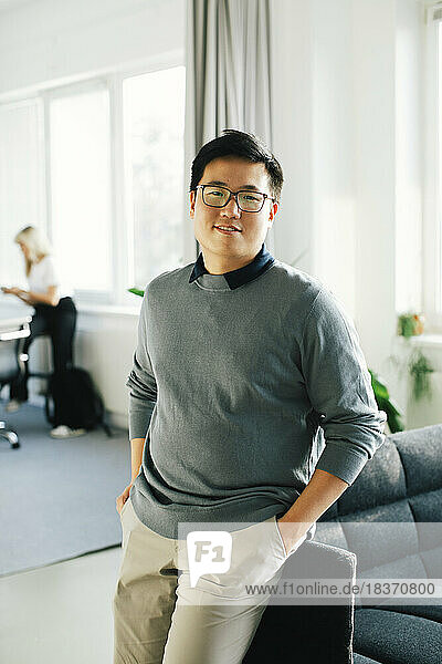 Porträt eines lächelnden jungen Geschäftsmannes mit Händen in den Taschen  der sich im Büro auf ein Sofa lehnt