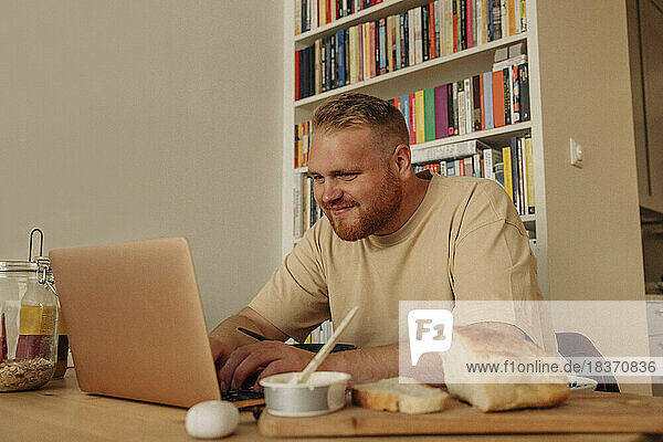 Lächelnder fettleibiger Mann  der einen Laptop benutzt  während er zu Hause auf dem Tisch sitzt und frühstückt
