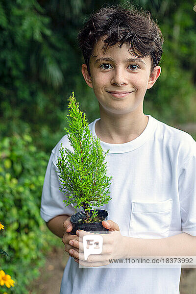 A boy holding a small tree sapling in a pot  standing in a garden.
