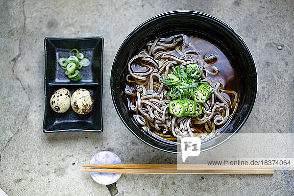 A black china bowl of noodles and broth  sliced green chilli and a dish with quail's eggs and sliced vegetables.